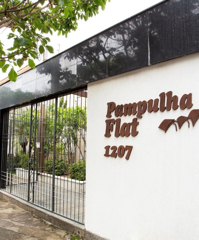 Visão da entrada do Pampulha Flat. Há um muro branco com o nome do local e o número 1207. Toda a entrada possui na parte de cima um mármore escuro e parte dela é de grade metálica com um pequeno jardim na parte interna do local.