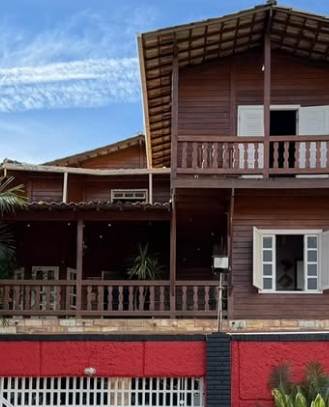 Visão da entrada do Woods Hostel BH. É uma casa de madeira de dois andares com um muro vermelho à frente. Em cada andar há uma janela branca. No primeiro, temos uma varanda ampla e no segundo uma pequena sacada. O local possui telhado de telhas coloniais.