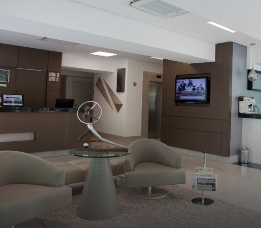 Imagem do Saguão do Bh Jaraguá Hotel. Há uma pequena mesa de centro com um escultura, algumas poltronas ao redor, balcão de atendimento ao fundo e na lateral direita uma televisão e a entrada do elevador.