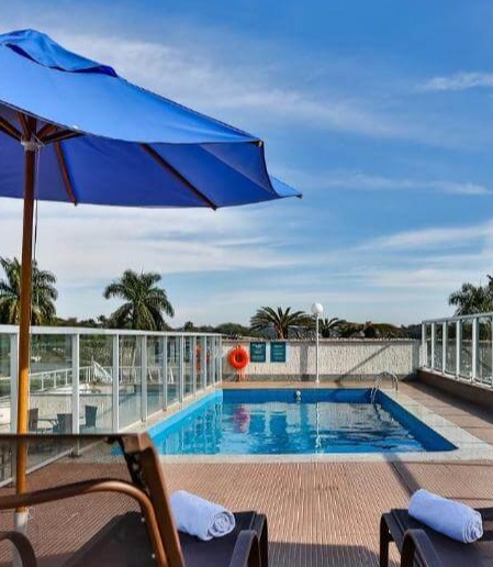 Imagem da área da piscina do hotel San Diego Suítes Pampulha. Há duas cadeiras com um guarda-sol azul e uma piscina cercada por grades de vidro.