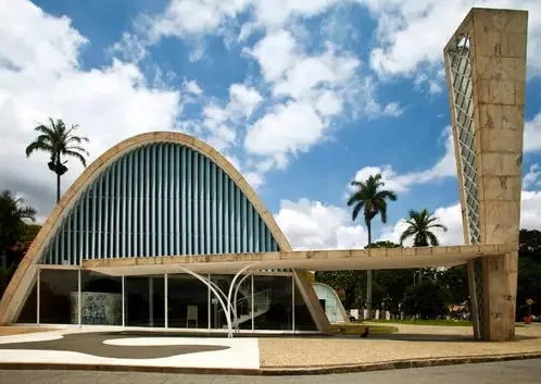 Entrada da Igreja da Pampulha, em Belo Horizonte, com fachada curva de concreto e vidro azul vertical, acompanhada de campanário alto à direita, céu azul com nuvens e palmeiras ao fundo.