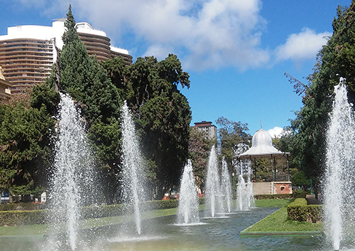 Chafarizes da Praça da Liberdade, em Belo Horizonte, com jatos de água formando arco e pequeno arco-íris sobre o espelho d’água, cercados por jardins e prédios históricos ao fundo.