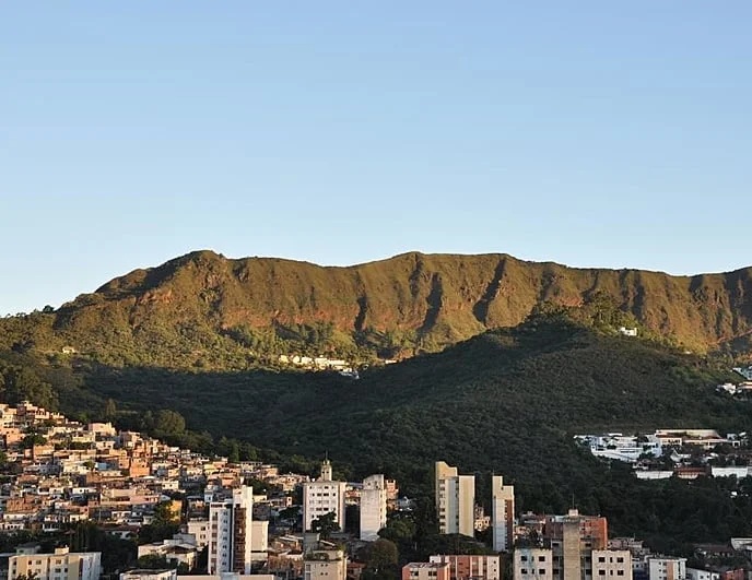 Visão do horizonte da cidade de Belo Horizonte em Minas Gerais. A imagem apresenta no centro um conjunto de montanhas, cercada por prédios da cidade e acima um céu azul sem nuvens.